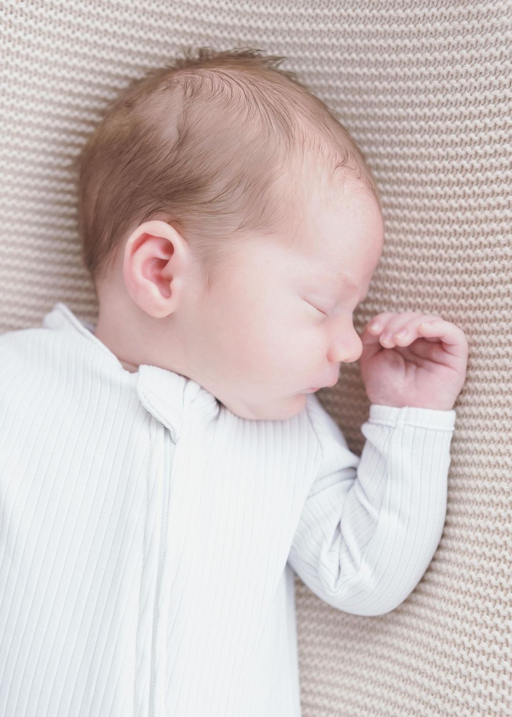 Peaceful newborn baby sleeping on a knit blanket, wearing a white outfit with hand raised to face.