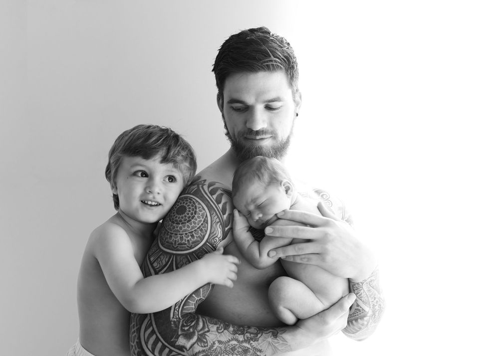 Black and white photo of tattooed father holding newborn baby while young son smiles beside him.