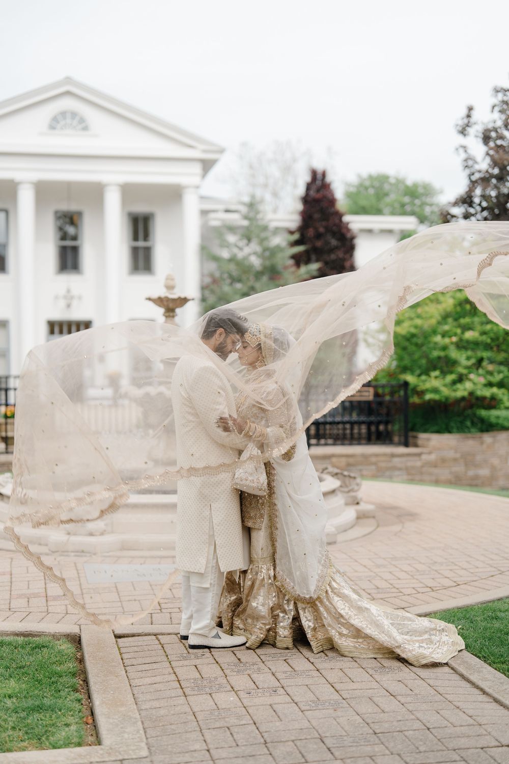 South Asian bride and groom in gold wedding attire share romantic moment under flowing veil at elegant white mansion.