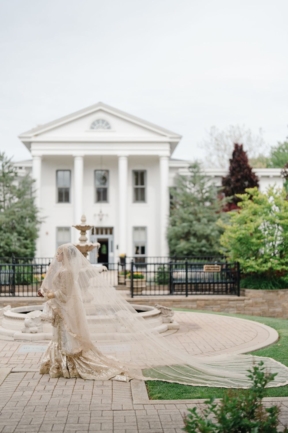 Bride in flowing gown and veil poses near fountain outside elegant white colonial wedding venue.