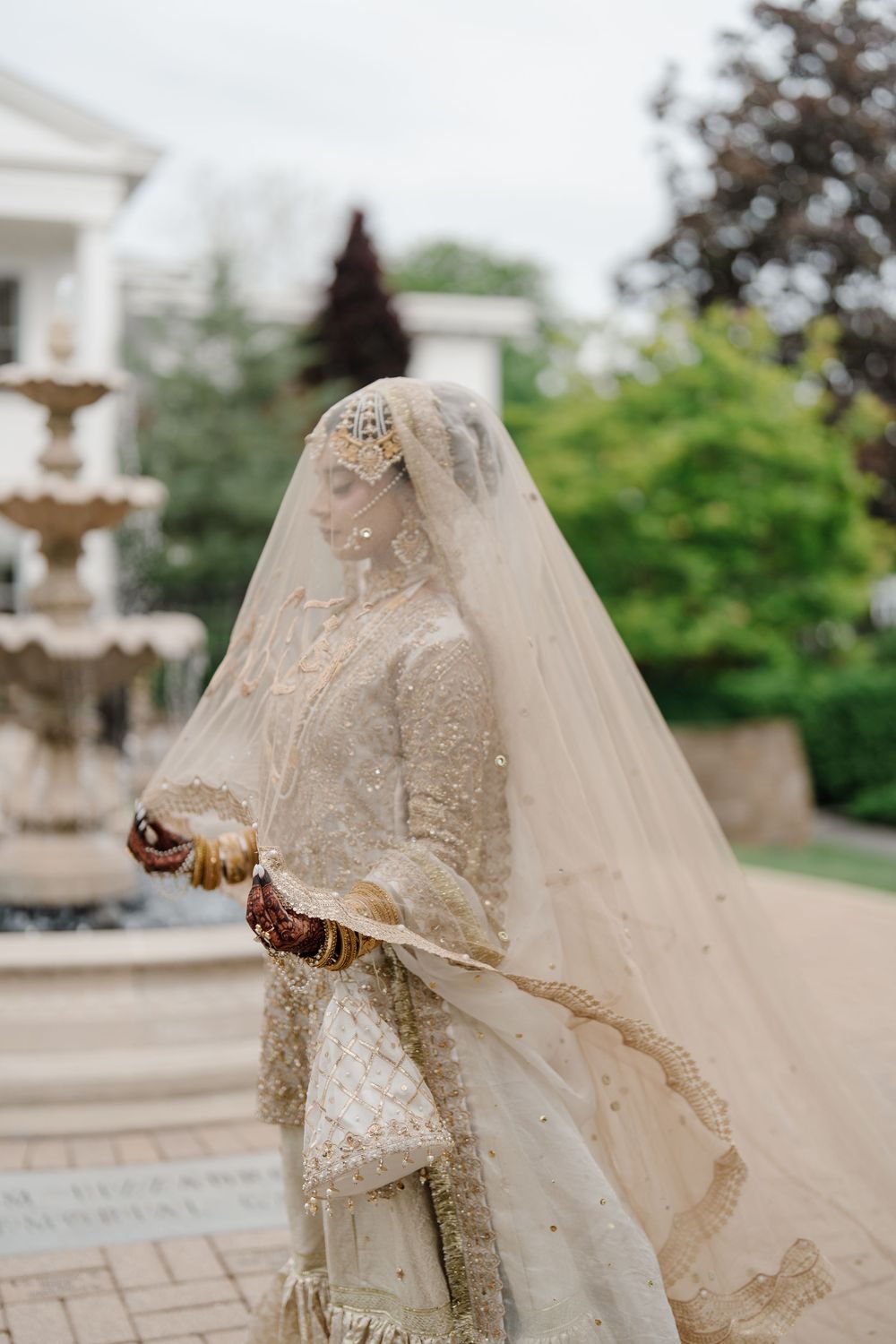 A bride in an ornate gold embroidered South Asian wedding dress and veil stands near a stone fountain in a lush garden.