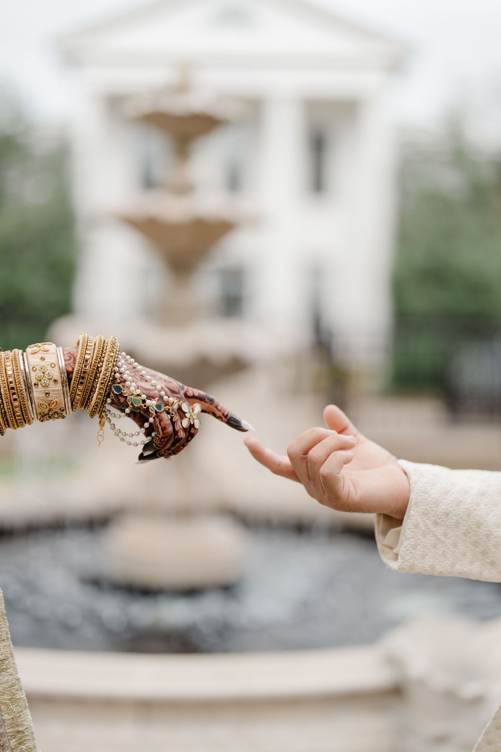Bride and groom touching fingertips, showcasing intricate henna and gold bangles, with a blurred fountain and white building backdrop.