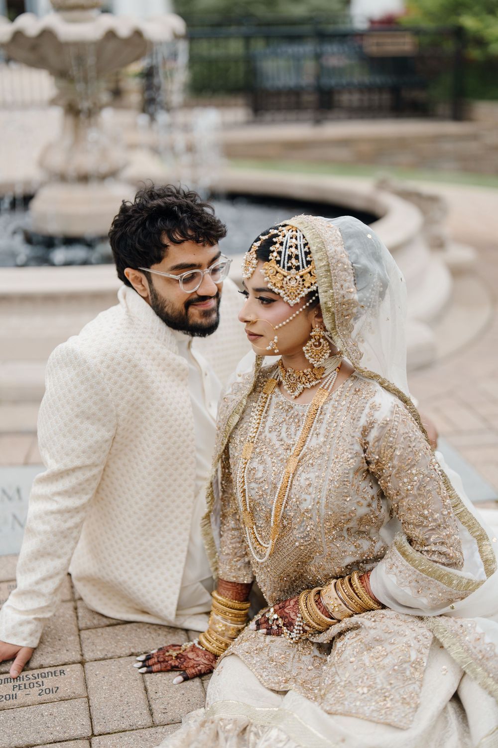 South Asian bride and groom in traditional gold and white wedding attire sit together near a fountain.
