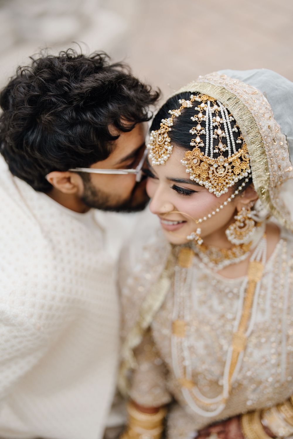 Groom in white sherwani kissing bride wearing ornate gold jewelry and embroidered dupatta at a South Asian wedding ceremony.