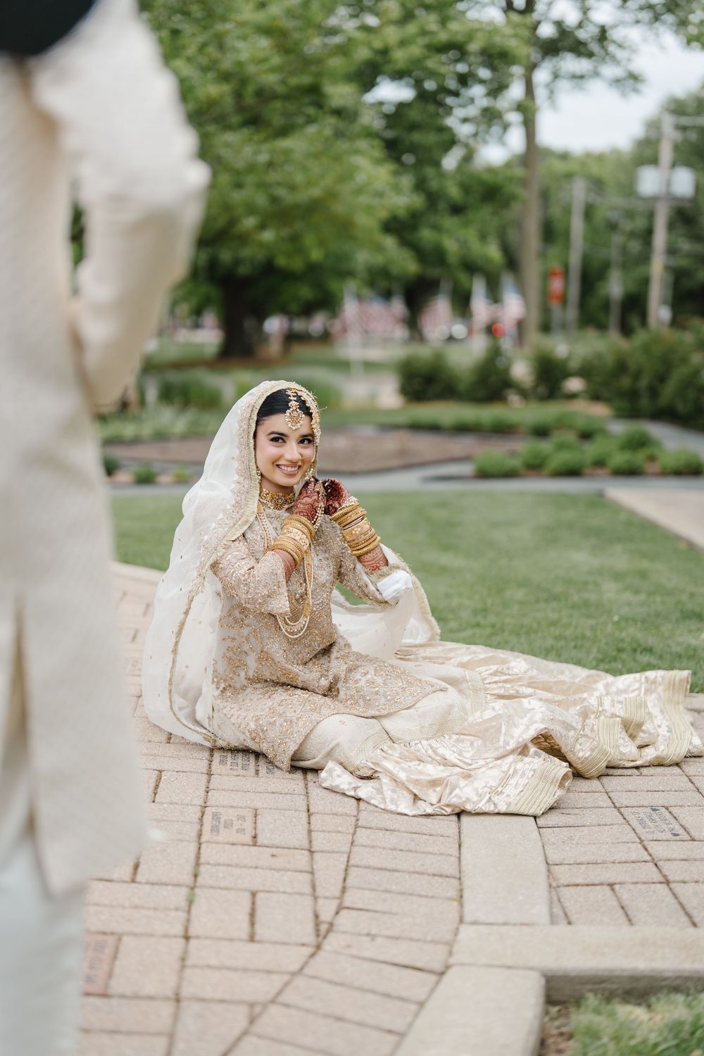 Smiling South Asian bride in ivory embroidered lehenga with gold jewelry and white dupatta sitting on brick pathway outdoors.
