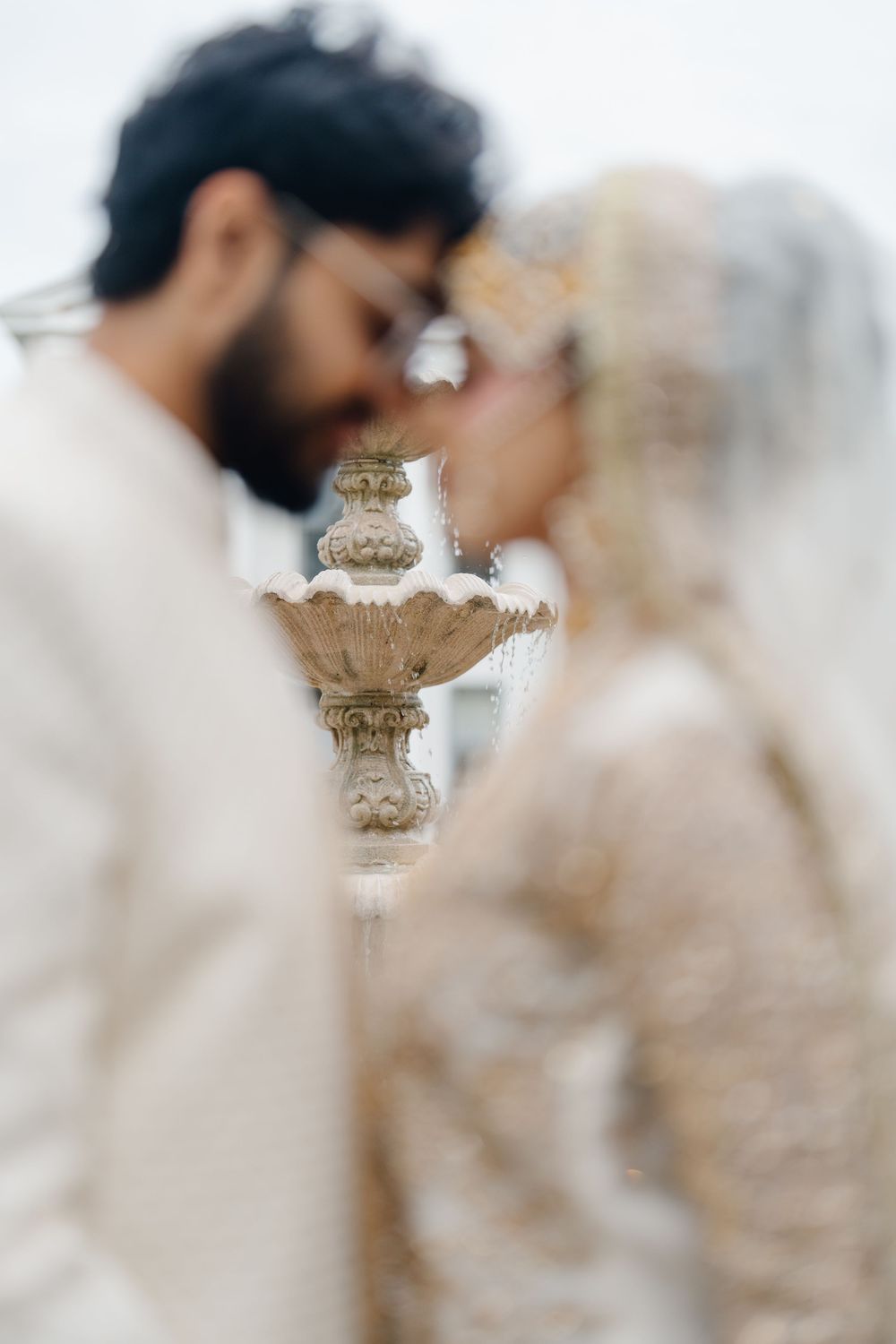 Couple in elegant white wedding attire sharing an intimate moment near an ornate stone fountain.