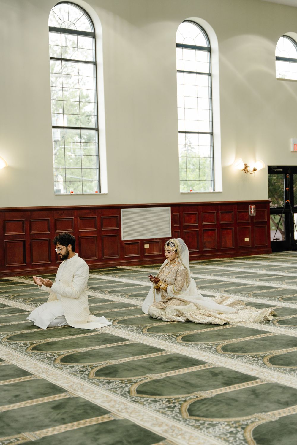 A couple in traditional white attire sits facing each other on a patterned carpet in a mosque with arched windows.