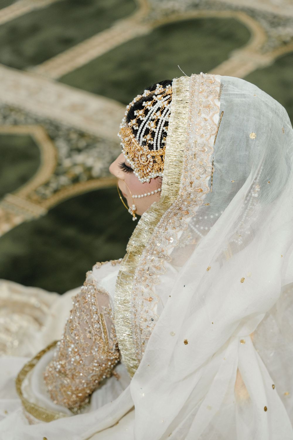 A bride in an ornate white and gold embroidered hijab with an elaborate pearl and gold crown headpiece smiles elegantly.