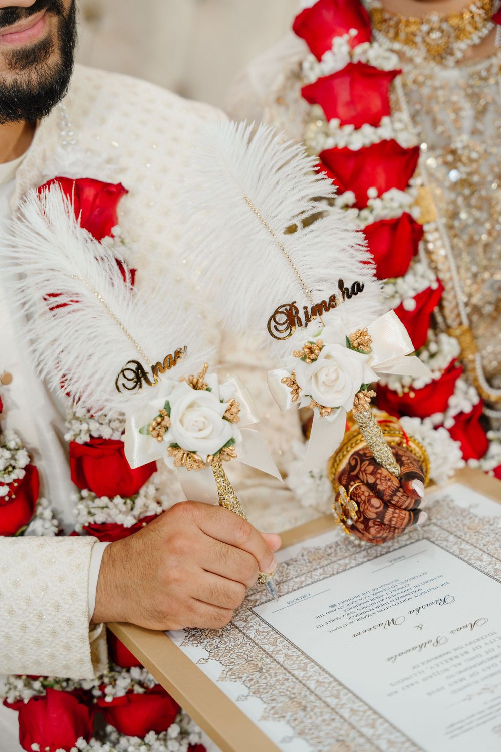 Indian wedding couple signing marriage certificate with ornate feather pens, adorned with red rose garlands and gold attire.