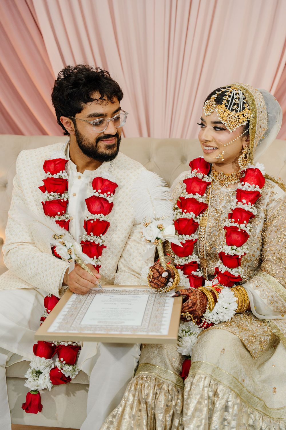 South Asian couple in white wedding attire with red rose garlands signing nikah marriage contract at ceremony.