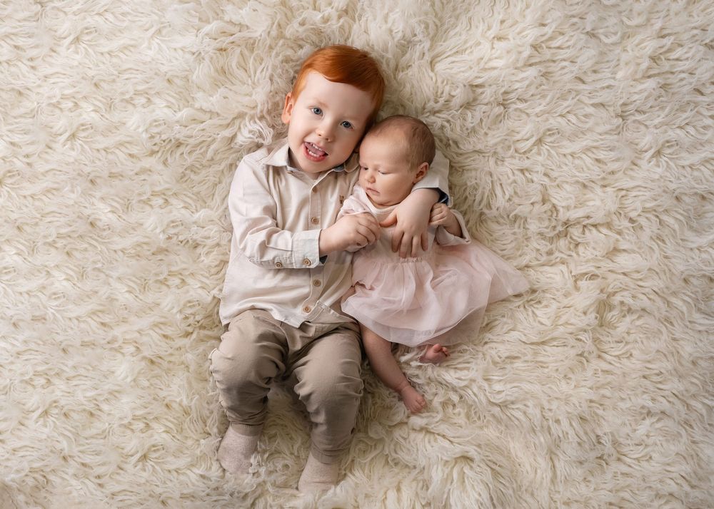 Two young children in neutral colored clothing lay on a plush white carpet while sharing sweet moments together.