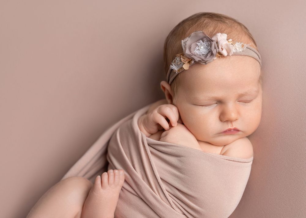 A sleeping newborn baby wrapped in soft beige fabric with a delicate lace headband during a professional photo session.