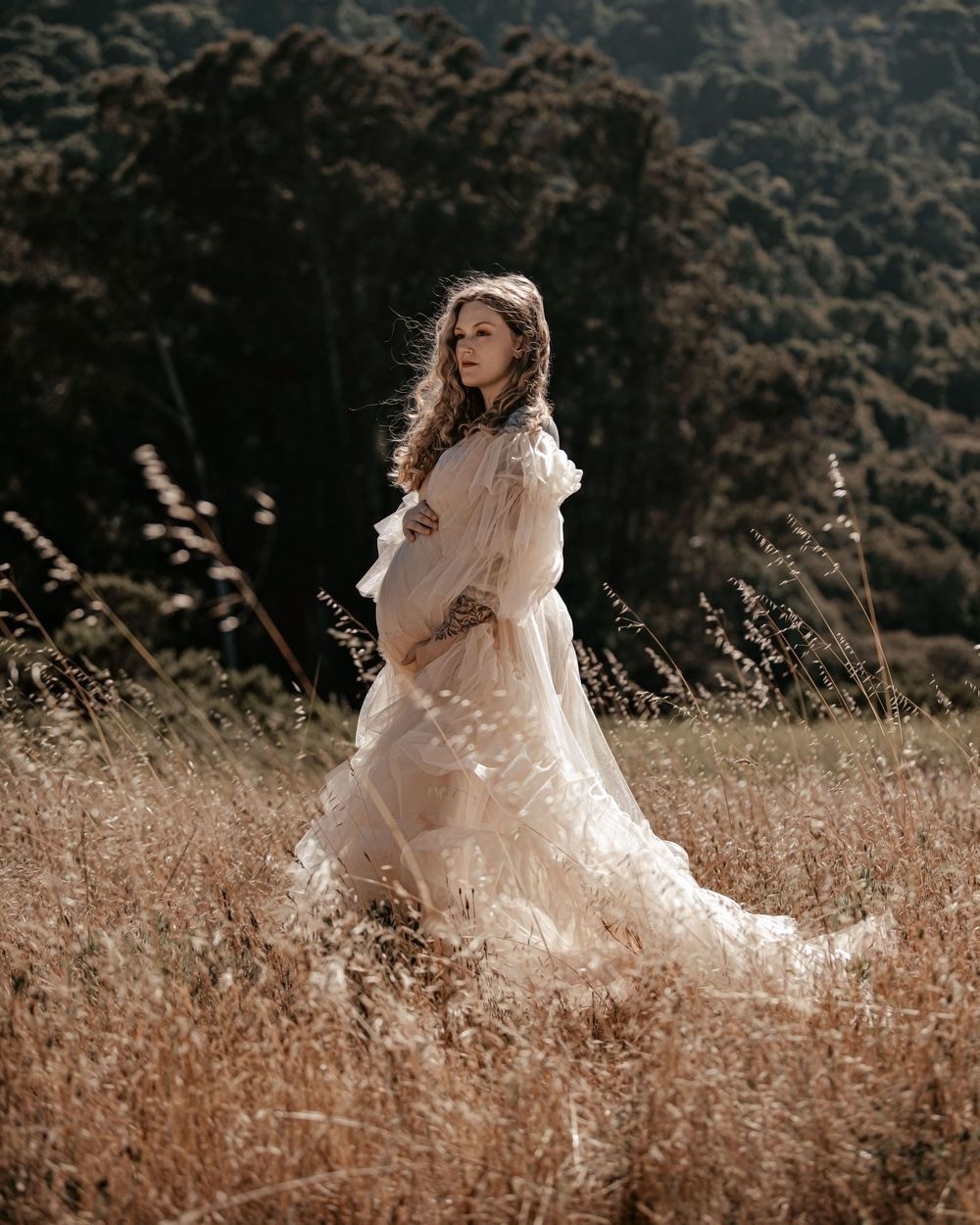 Pregnant woman in flowing white lace gown standing gracefully in golden meadow with lush green hills backdrop.