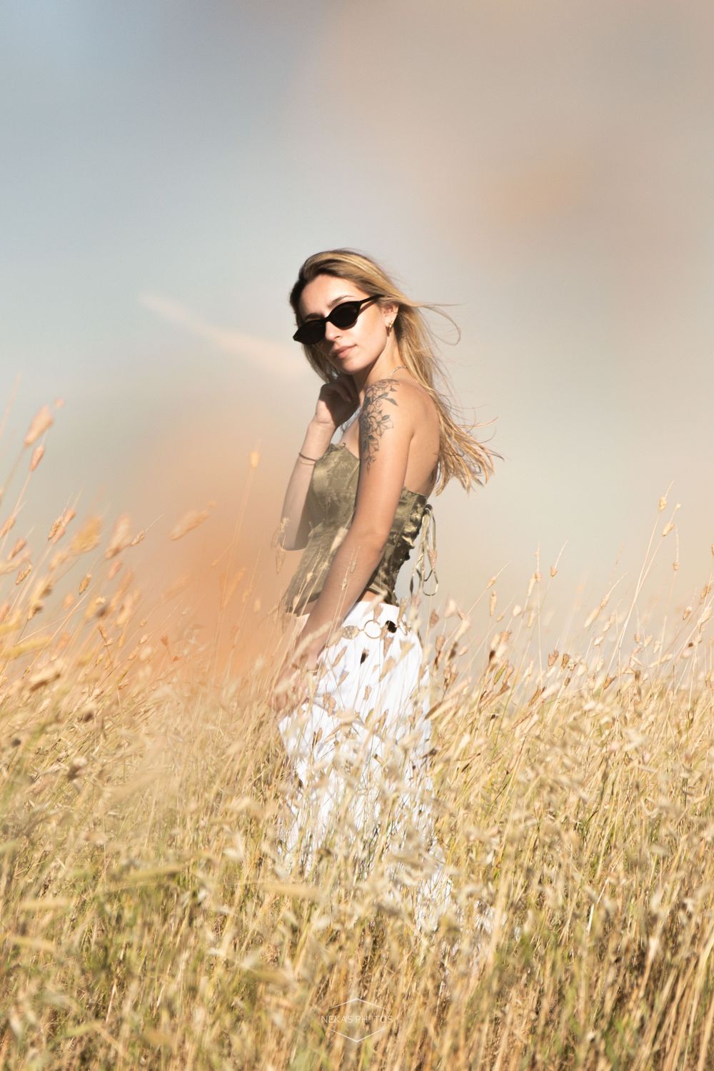 Blonde woman in sunglasses and white dress standing in golden wheat field with dreamy sky background.