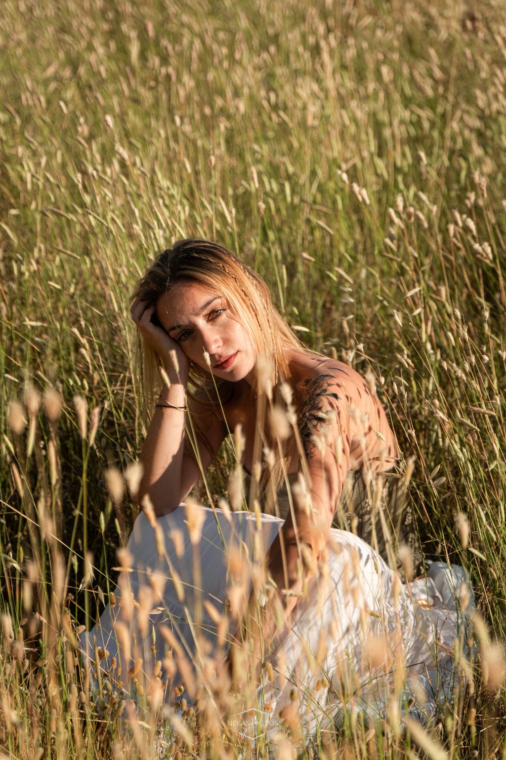 Young woman with long hair sitting in tall golden grass field, wearing floral top and white skirt in warm sunlight.
