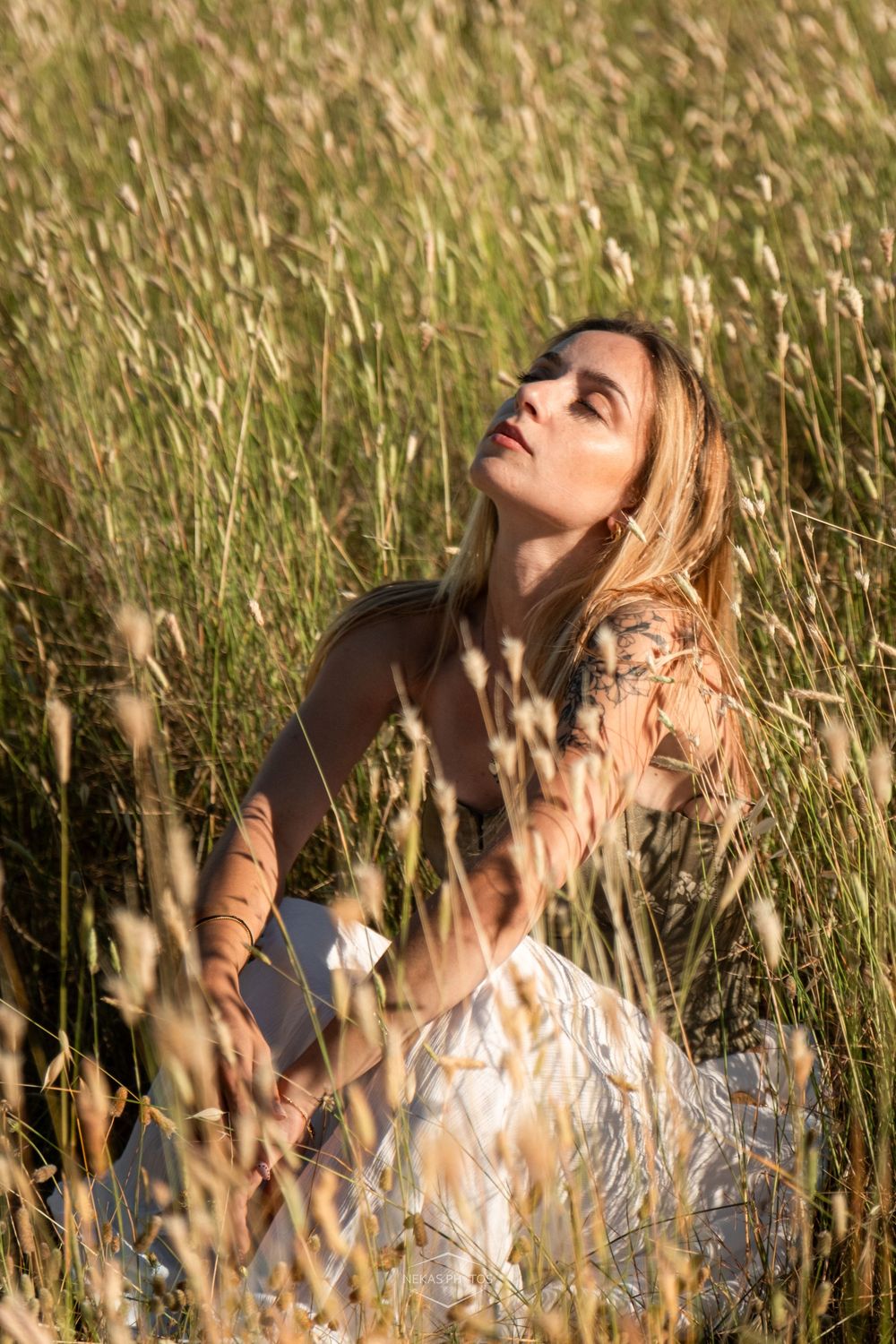 Woman with tattoos sitting in tall golden grass field, wearing white dress, looking up in warm sunlight.
