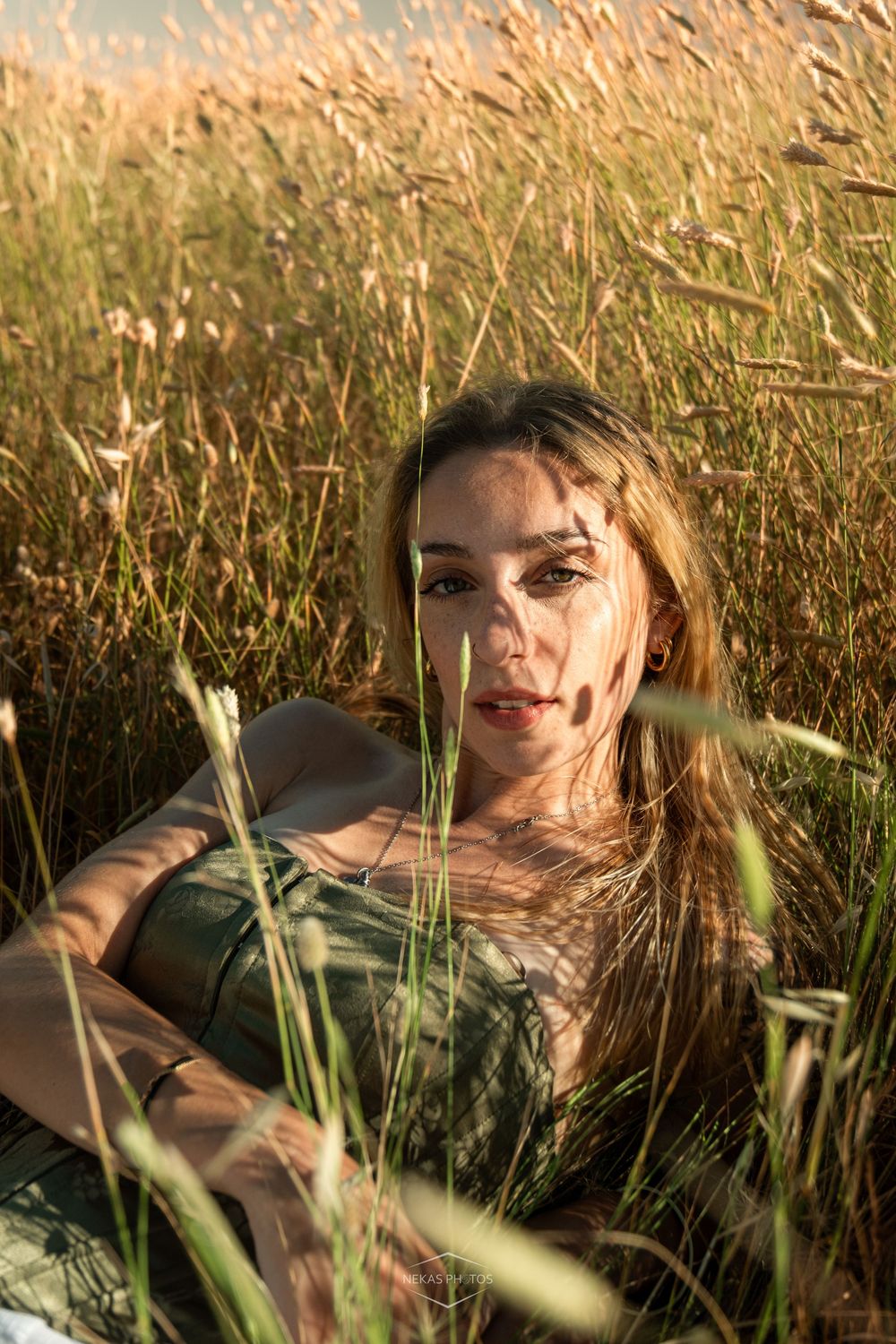 Young woman with long hair sitting in golden tall grass field, bathed in warm sunset light, gazing at camera.