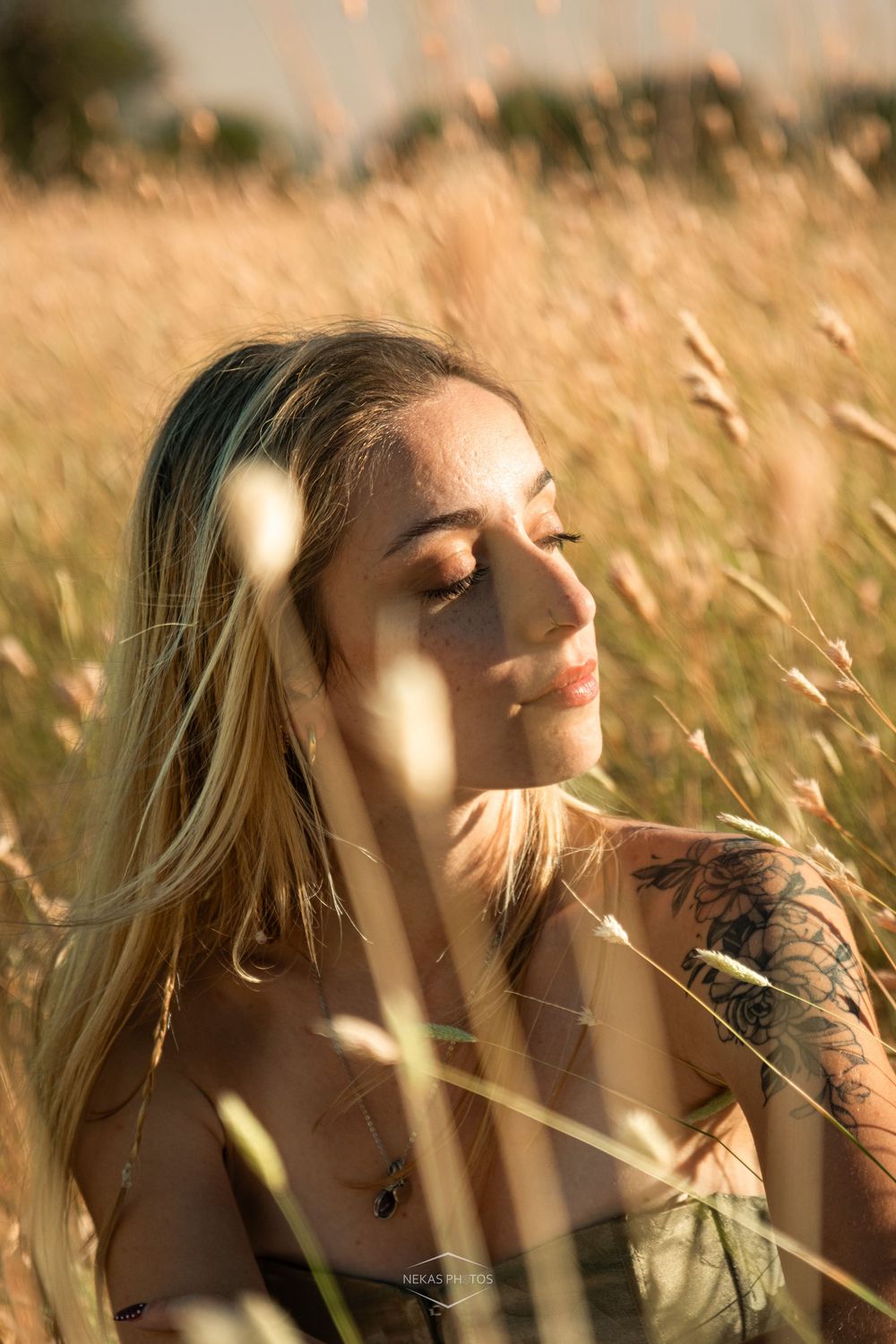 Blonde tattooed woman with closed eyes sitting peacefully in a golden wheat field during golden hour sunlight.