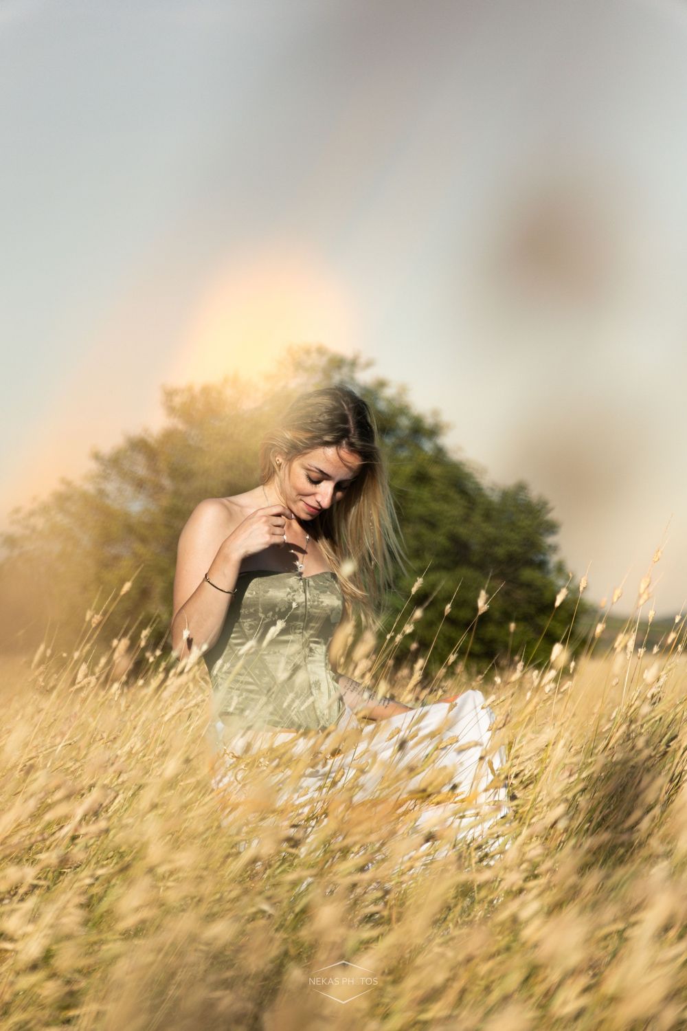 Young woman in strapless dress sitting in golden tall grass field during sunset with warm glowing light.