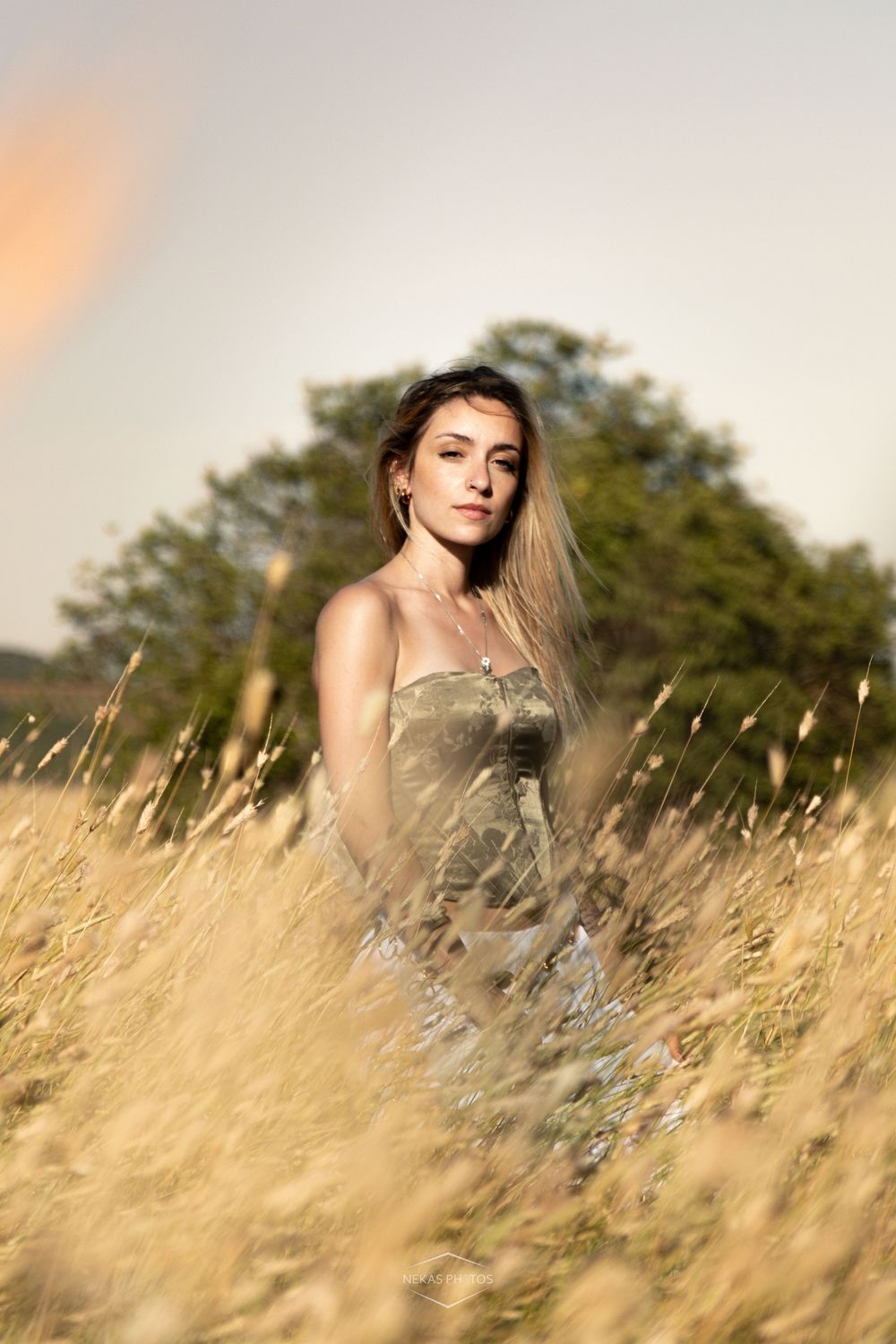 Young woman in olive dress standing in golden tall grass field at sunset with trees in background.