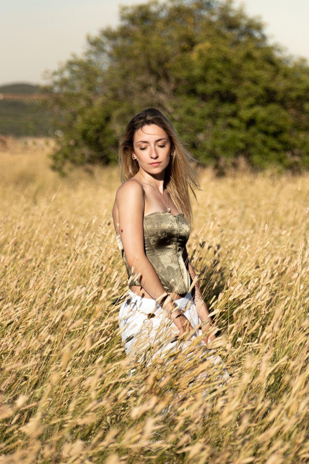 Young woman in floral dress sitting peacefully in golden wheat field with trees in background on sunny day.