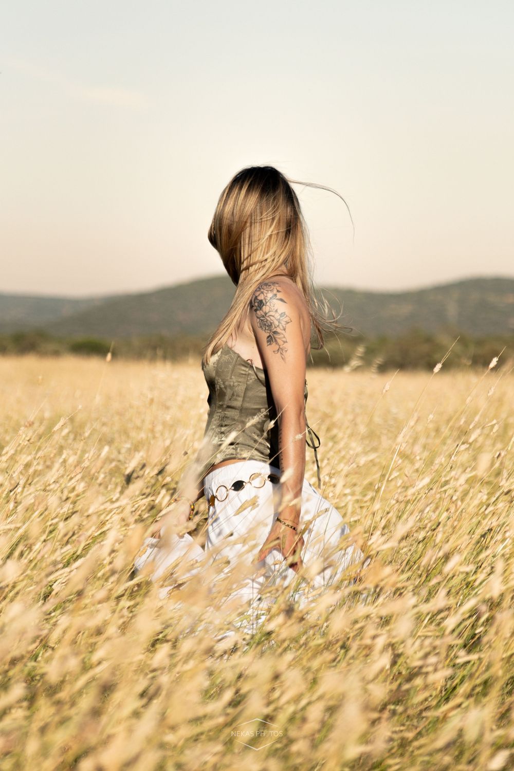 Blonde woman with tattoo in olive top and white skirt standing in golden wheat field with hills in background.