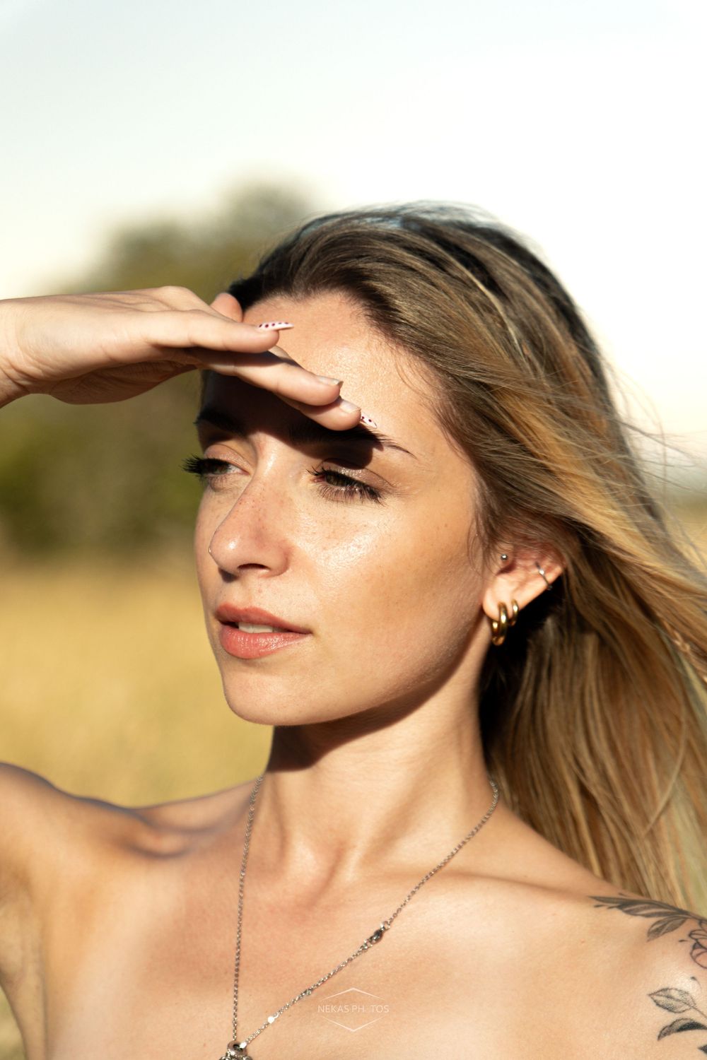 Young woman with tattoos shielding her eyes from the sun in a golden field, wearing silver jewelry.