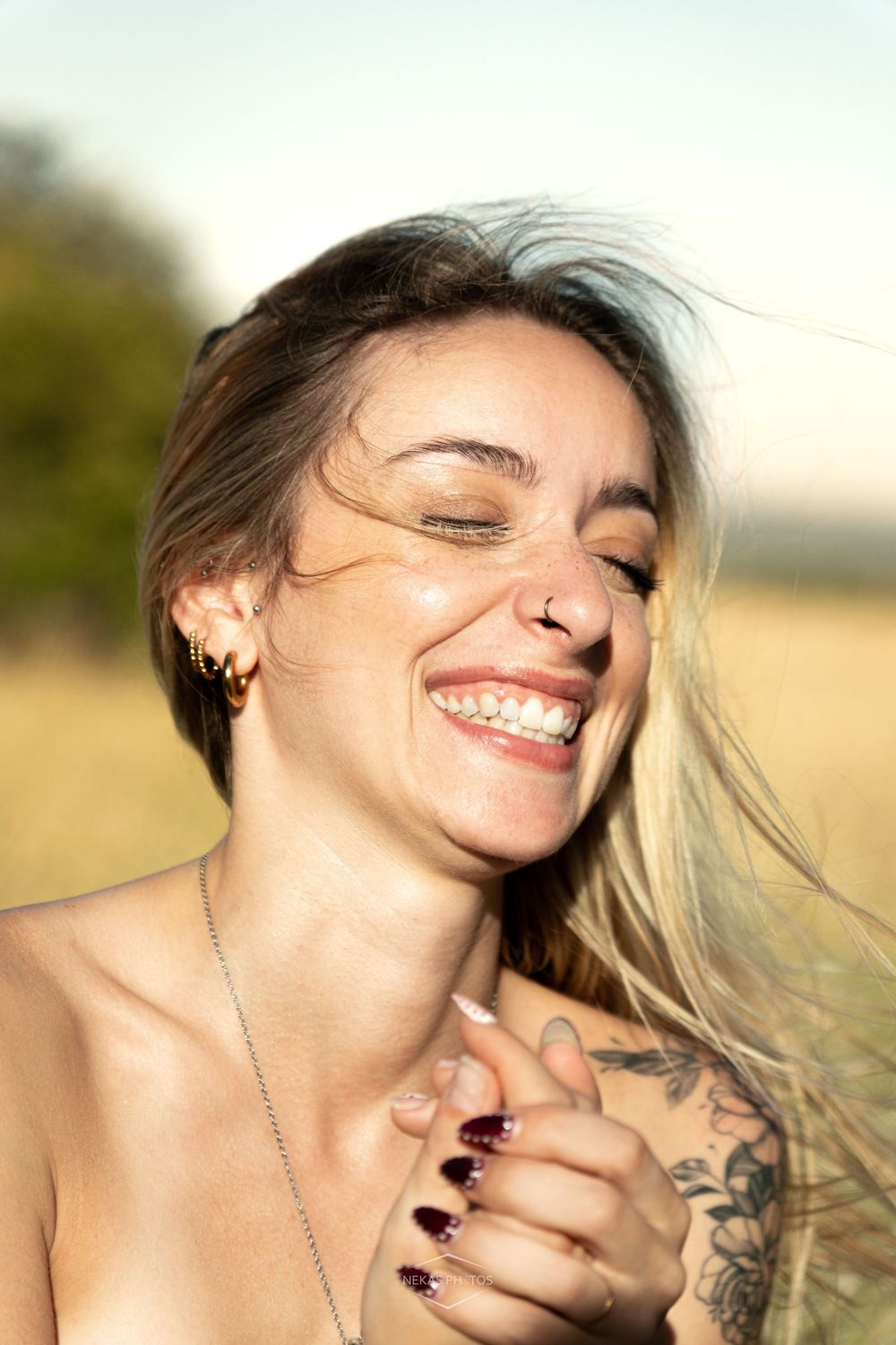Joyful woman with tattoos, nose ring, and windswept hair laughing outdoors in a sunny field.