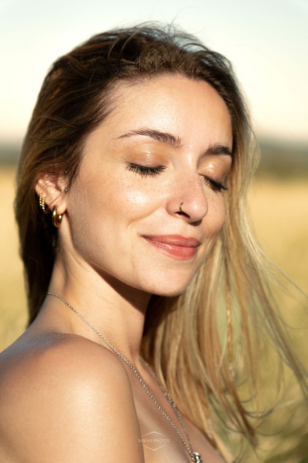 Young woman with freckles, nose ring, and long hair smiles serenely with closed eyes in golden sunlight outdoors.