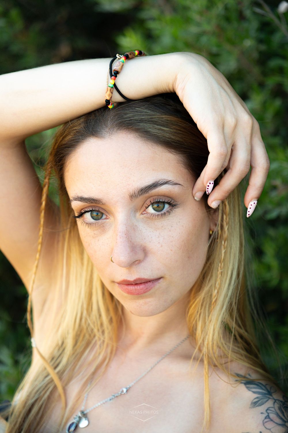 Young woman with freckles, green eyes, colorful bracelet, and painted nails poses outdoors with hand on head.