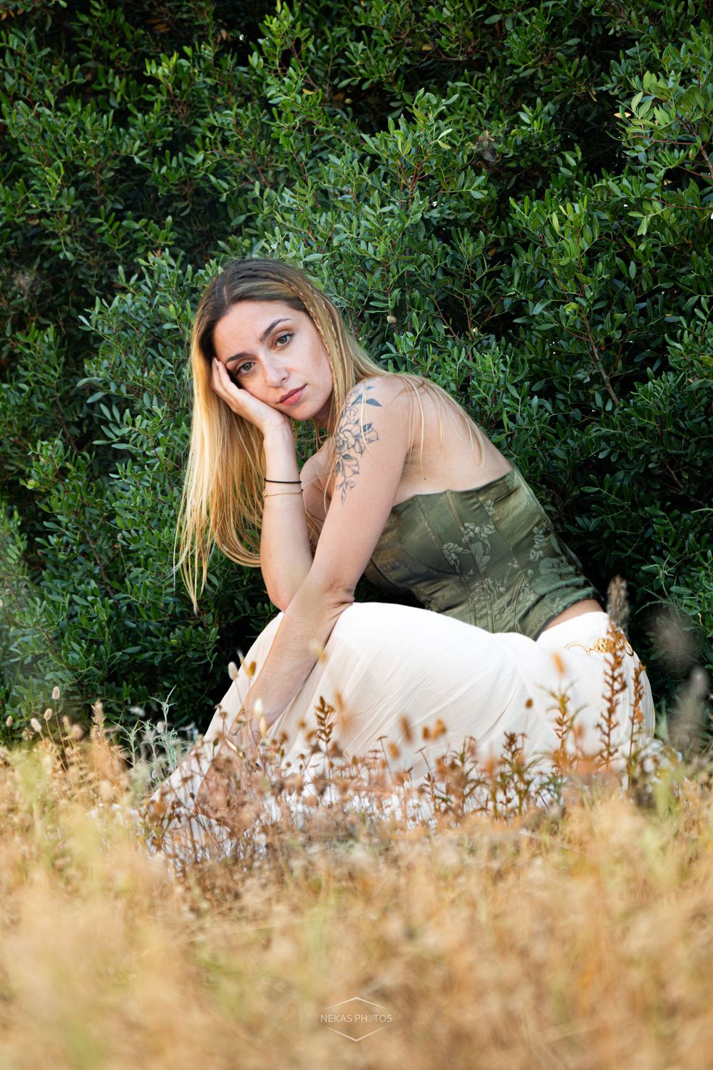Young woman with tattoos sits in golden grass before lush green bushes, wearing olive top and white skirt.