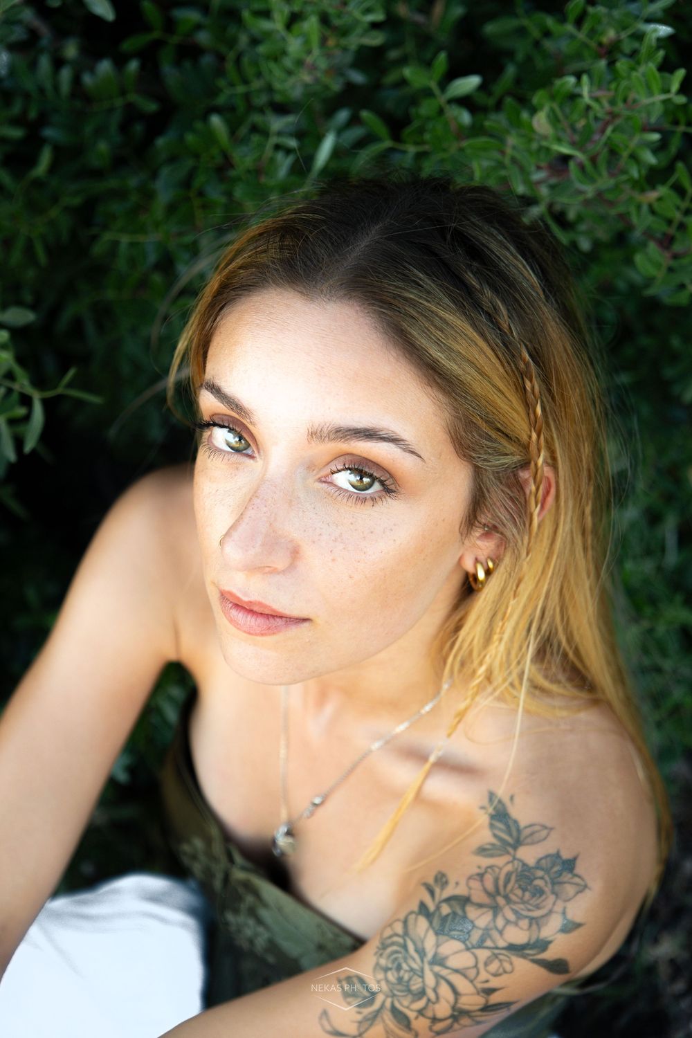 Young woman with freckles, braided hair, and floral arm tattoo poses outdoors against lush green foliage.