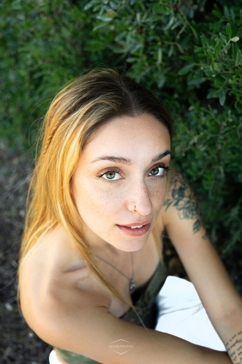Young woman with freckles and nose ring posing outdoors near green foliage, looking up at camera.