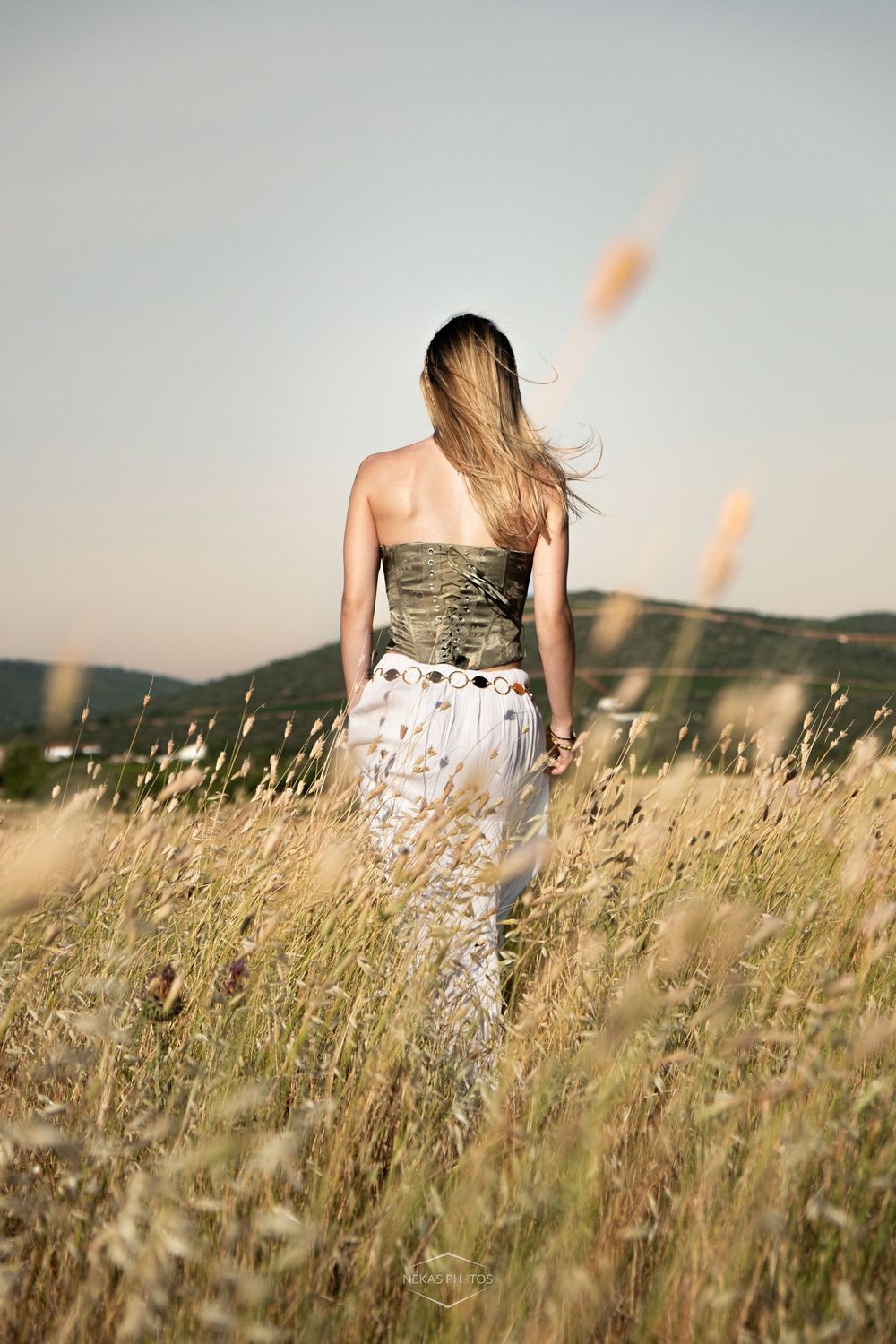 Woman in strapless top and white skirt walking through golden field with hills in background.
