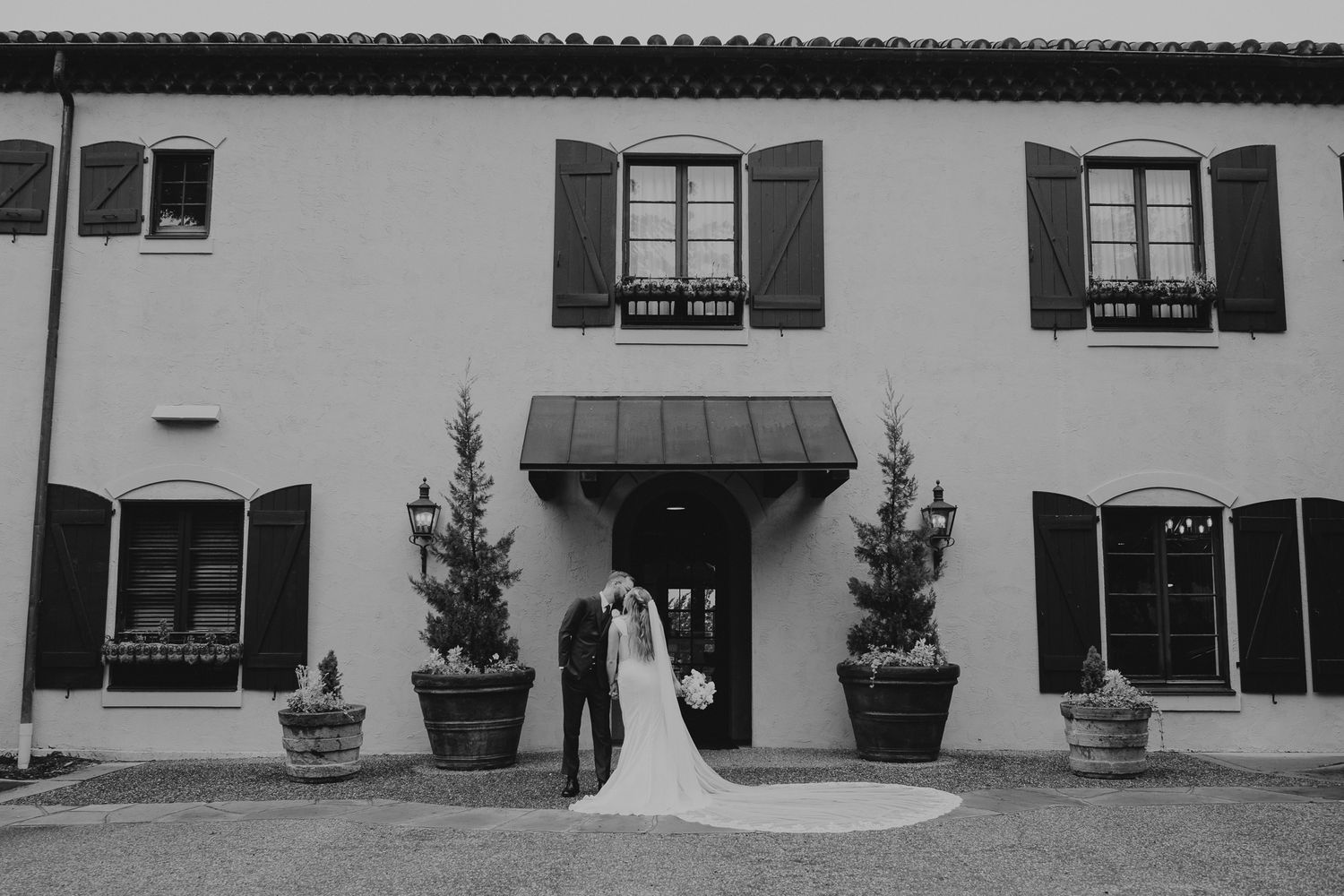 Black and white photo of a bride and groom walking through a tree-lined path, groom holding the brides flowing gown.