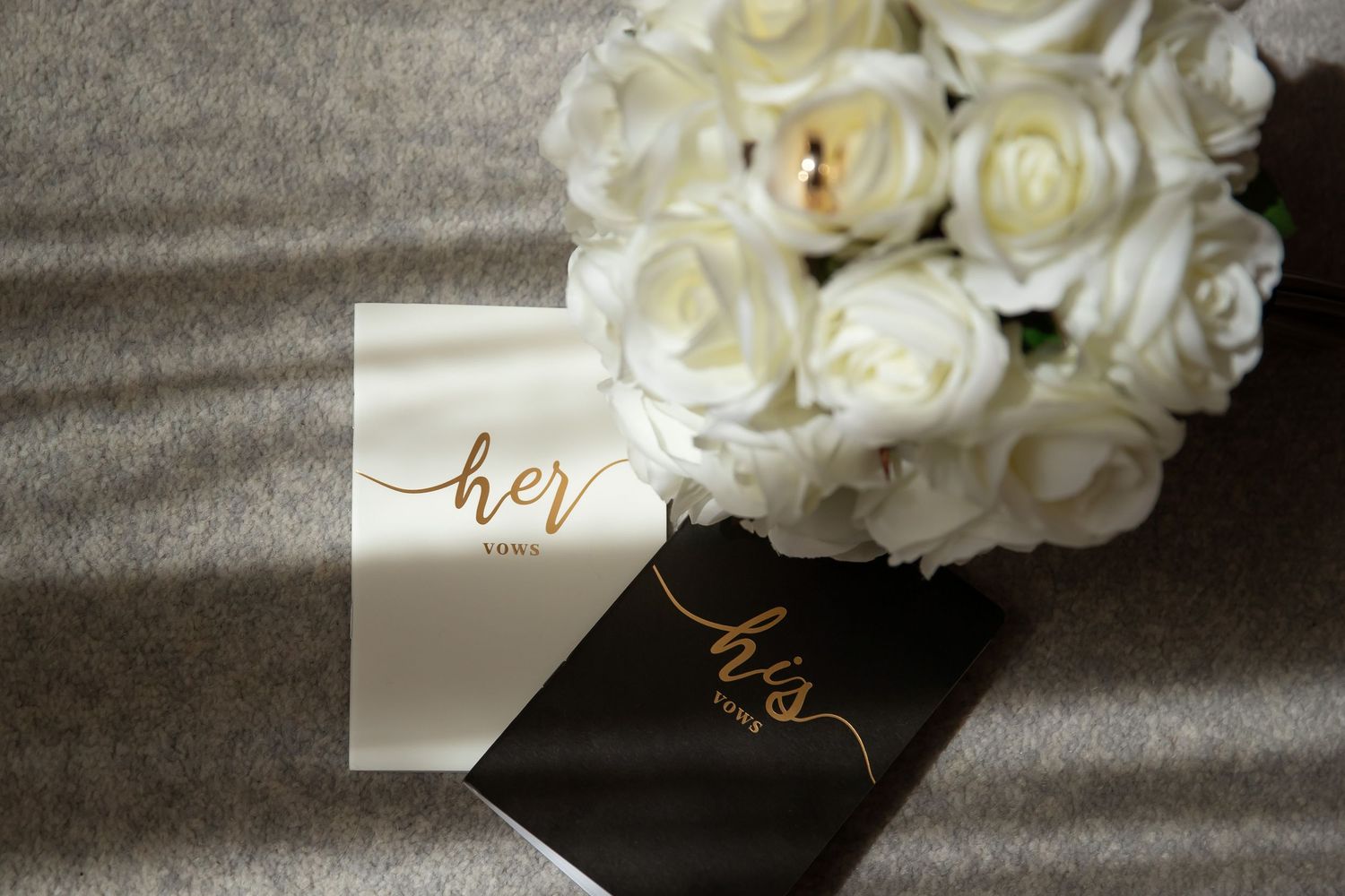 White rose bouquet beside gold script his and her wedding vow books on a gray surface.