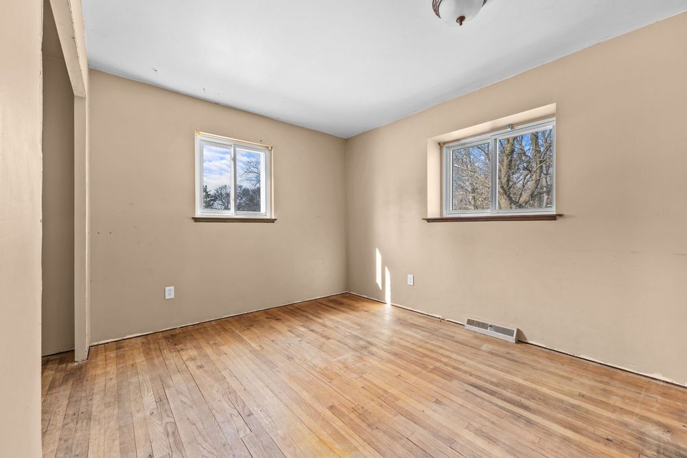 Sunlit empty bedroom with hardwood floors, beige walls, two windows, and a ceiling light fixture.