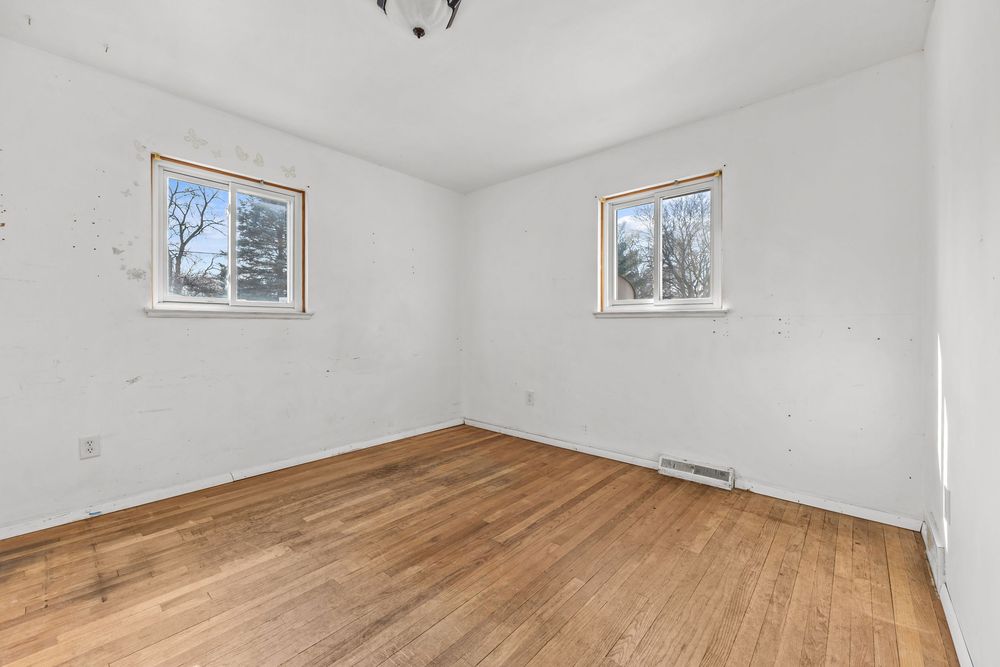 Vacant bedroom featuring hardwood floors, white walls, two windows, and a baseboard heater.
