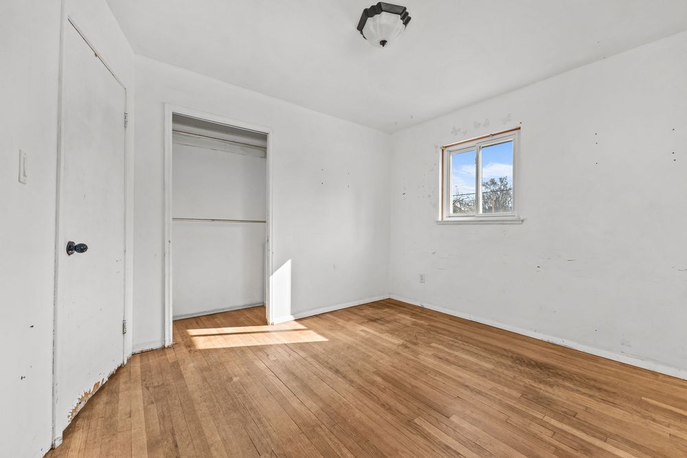 Empty bedroom with hardwood floors, white walls, open closet, and a bright window.