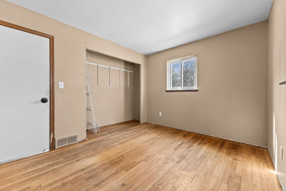 Vacant bedroom with hardwood floors, beige walls, open closet with curtain rod, and a window.