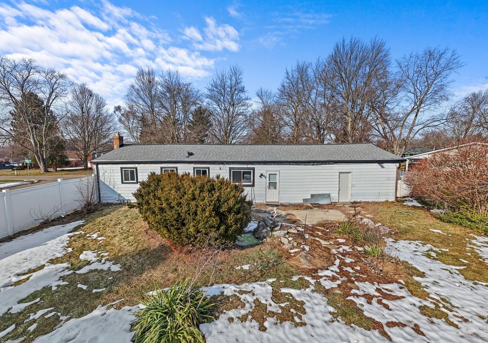 Ranch-style home exterior in winter with snow-covered yard, shrubs, and bare trees in background.
