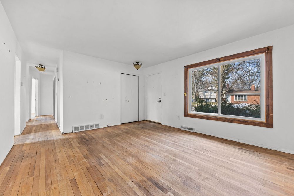 Bright empty living room with hardwood floors, white walls, and a large window overlooking trees.