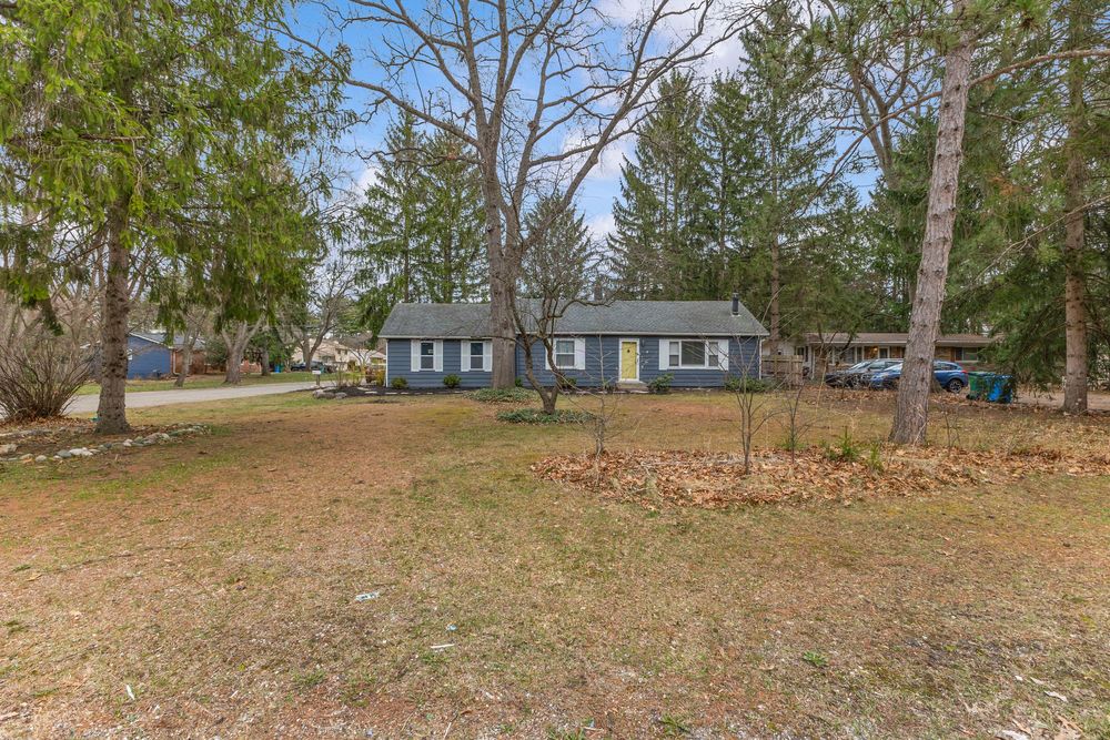 Blue ranch-style home surrounded by mature trees and a wide open front yard in early spring.
