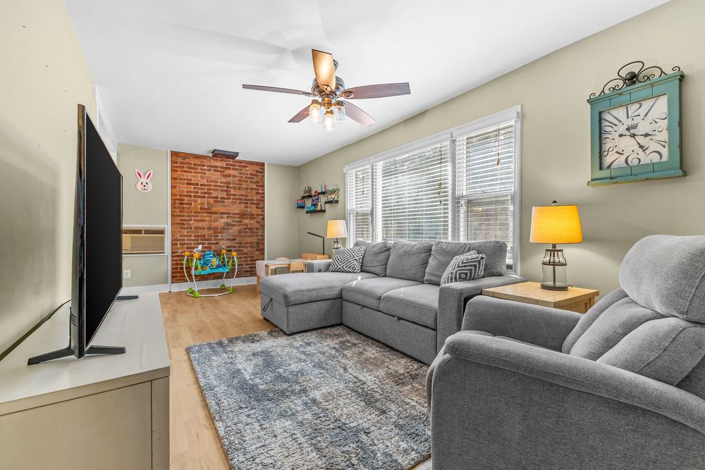 Cozy living room featuring gray sofas, exposed brick accent wall, ceiling fan, and hardwood floors.