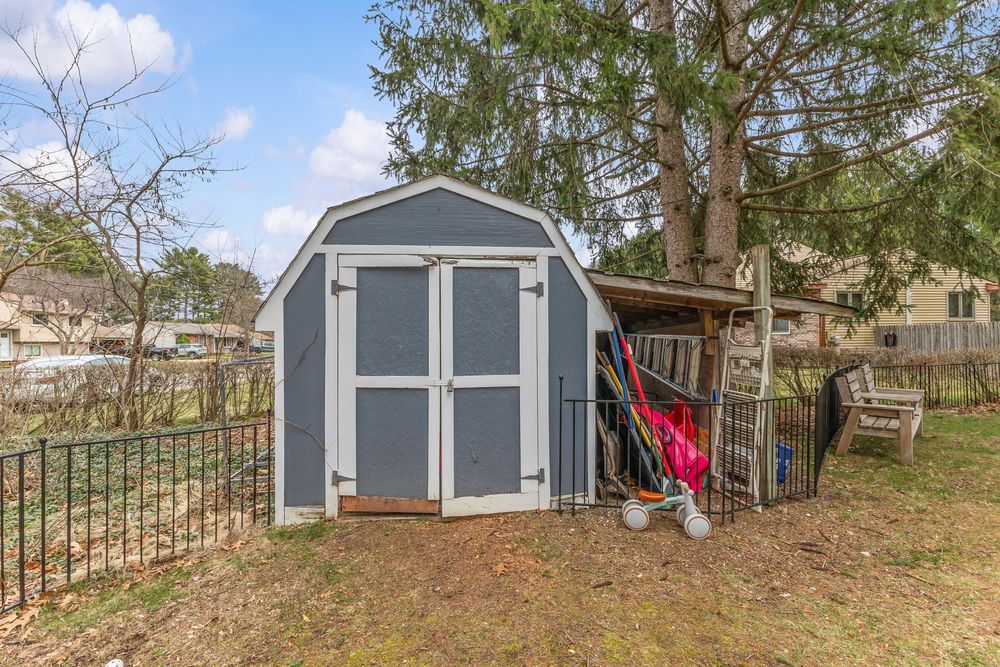 Gray storage shed with barn-style roof in a fenced backyard with mature trees and a wooden bench.