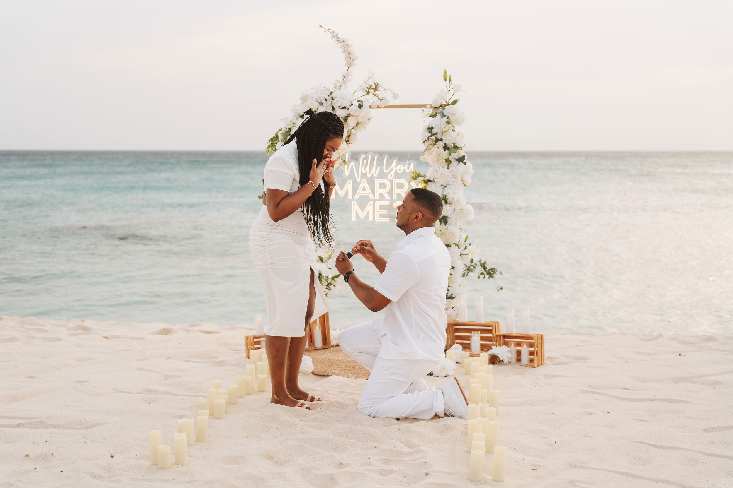 Man proposes to woman on a romantic beach setup with candles, floral arch, and Will You Marry Me sign.
