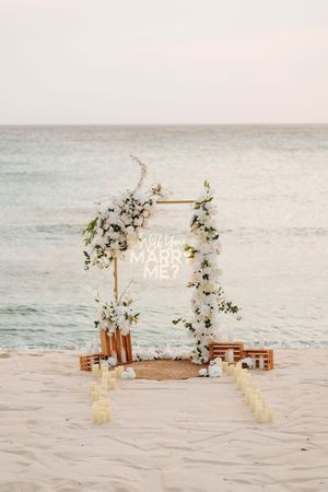 Beach proposal setup with floral arch, neon Will You Marry Me sign, candles on white sand, ocean backdrop at sunset.