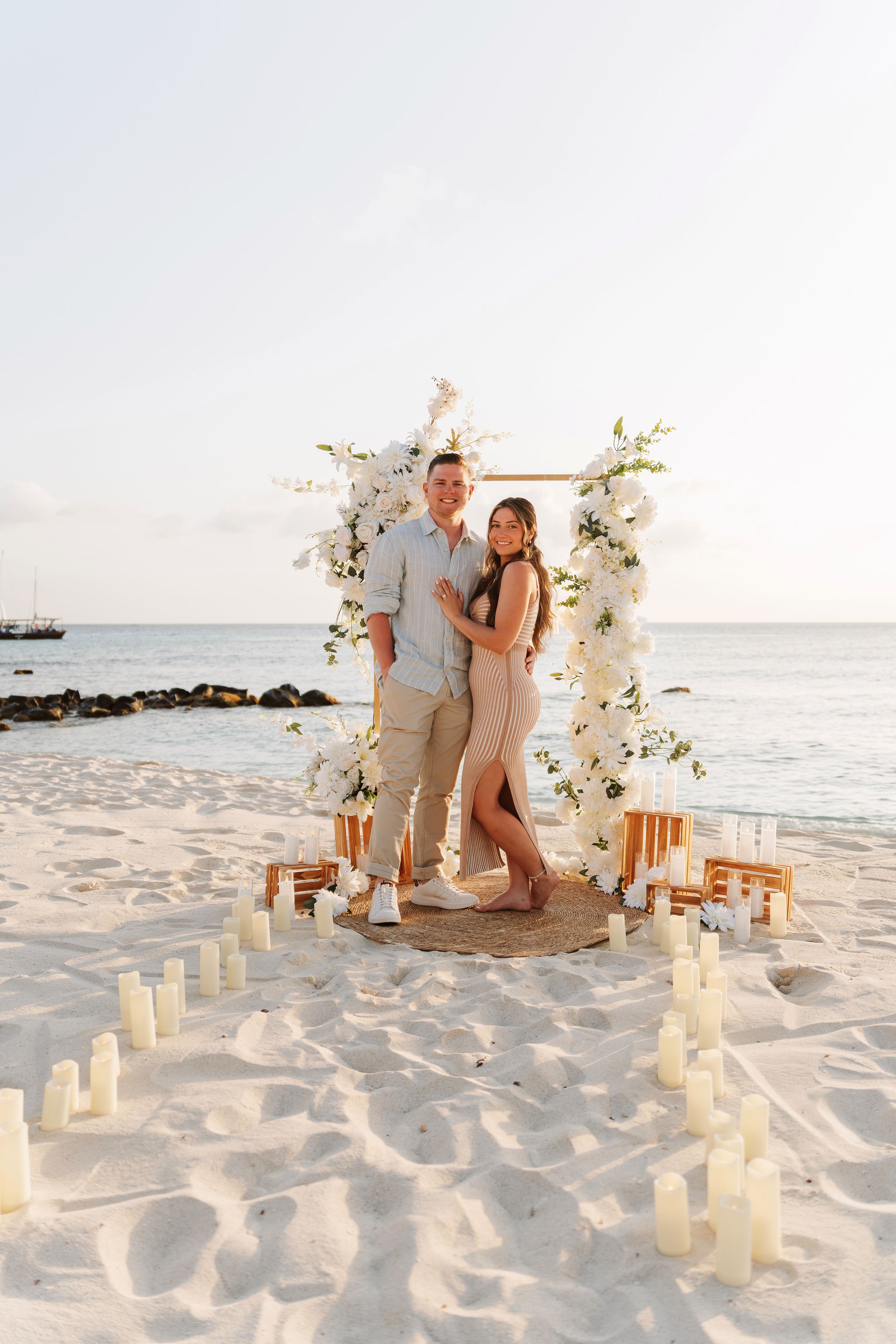Couple poses at beach proposal setup with white floral arch, candles in sand, and ocean backdrop at sunset.