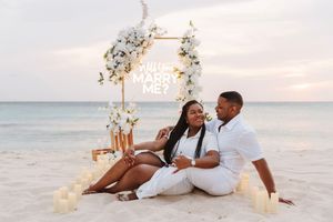 Couple sits on beach at sunset near a floral arch with a Will You Marry Me sign surrounded by candles.