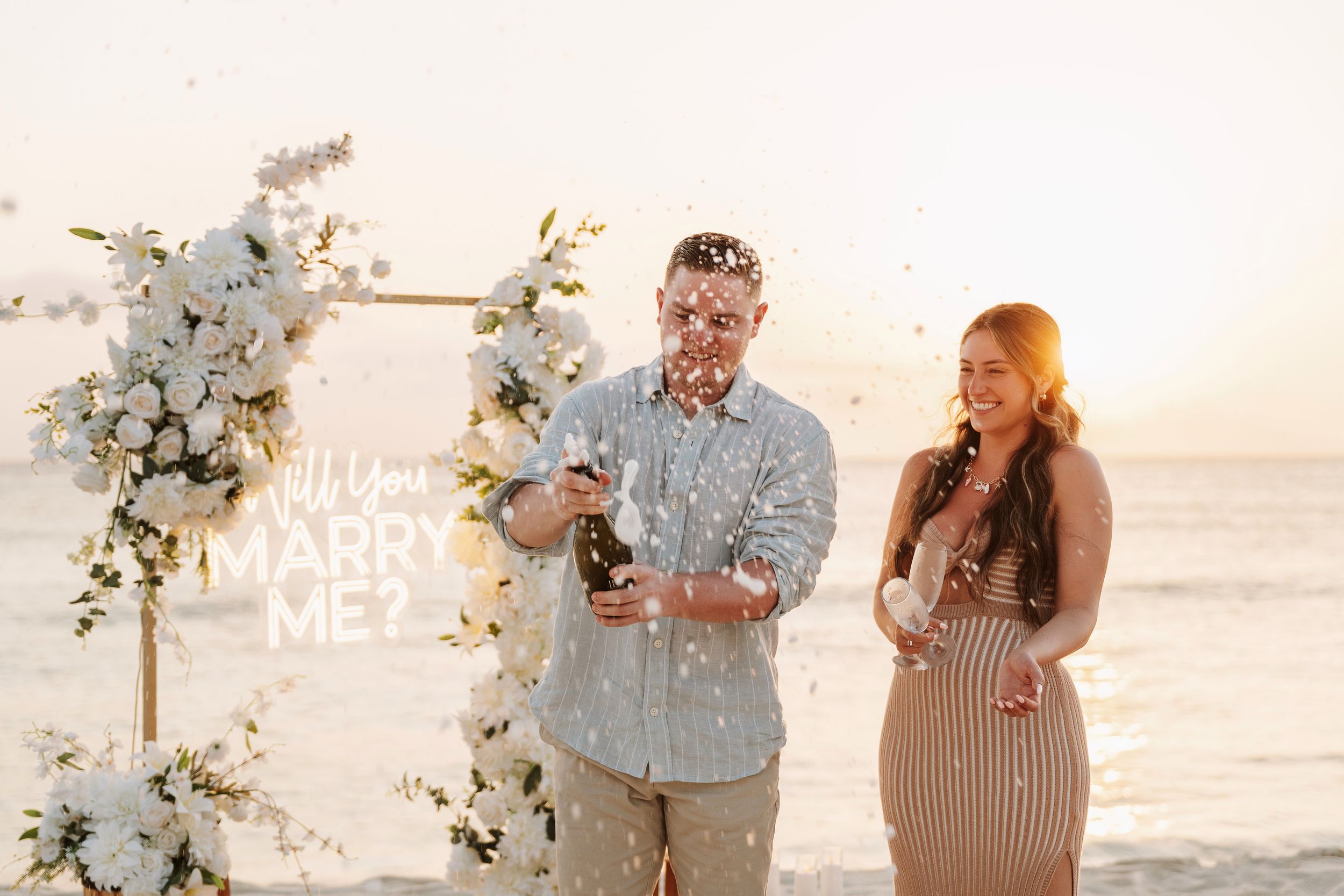 Couple celebrating beach proposal with confetti, champagne, and floral arch with Will You Marry Me sign at sunset.